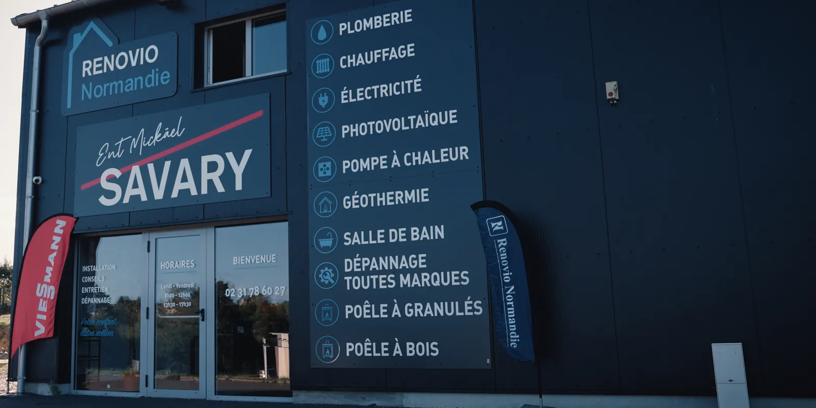 Façade en bardage noir du bureau d’Aunay-sur-Odon, avec l’enseigne Renovio Normandie, l’enseigne SAVARY et la colonne des prestations, de la plomberie au poêle à bois.