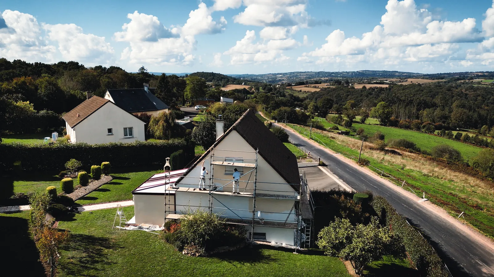 Vue du ciel d’une maison au pignon sous échafaudage, deux peintres au travail, au milieu du bocage normand.