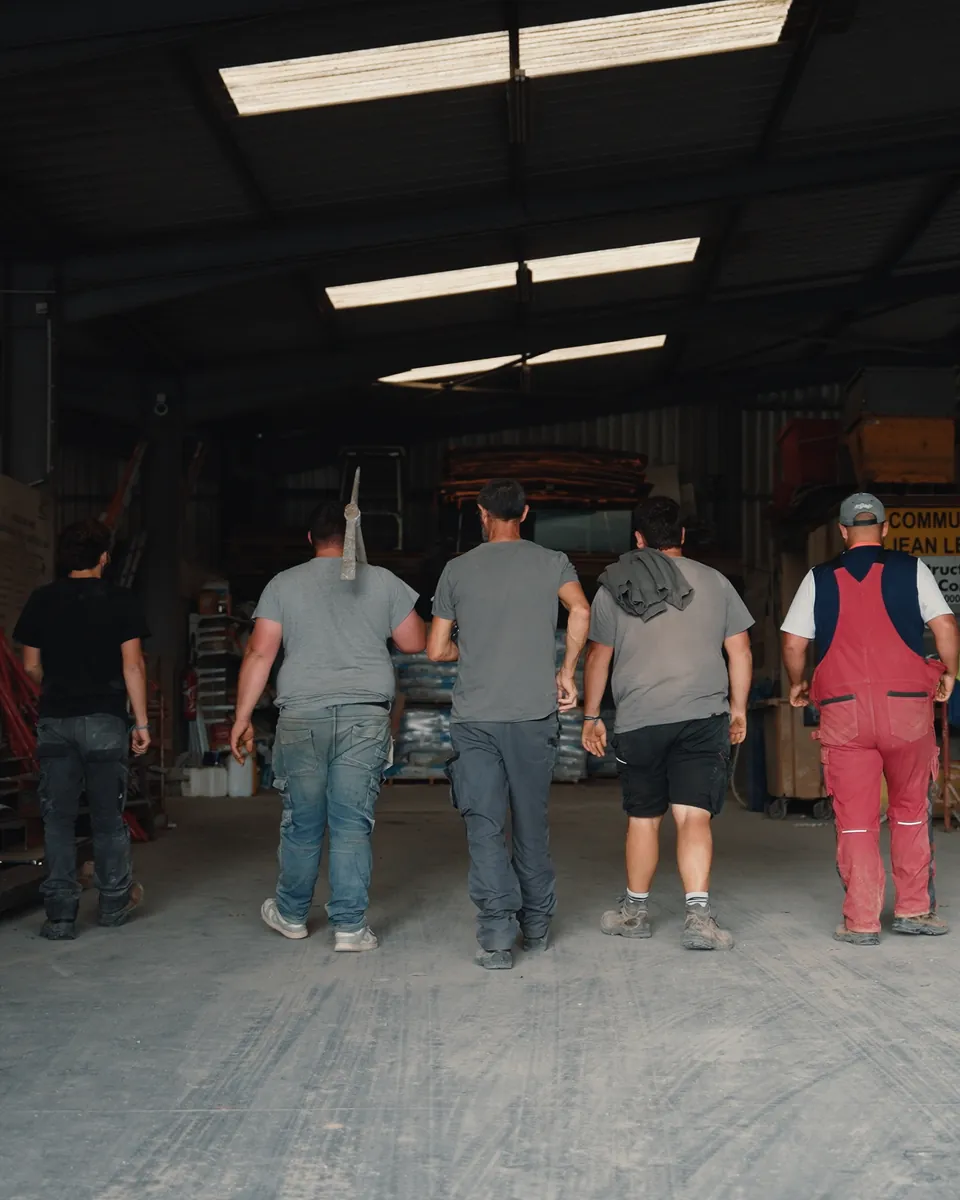 Quatre ouvriers, vus de dos, entrent ensemble dans un hangar de stockage de matériaux.