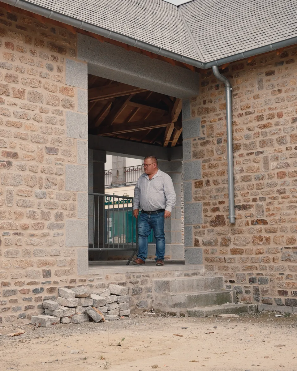 Un homme debout dans l’ouverture d’un bâtiment en pierre : murs en moellons, chaînes d’angle et encadrement en granit, marches de pierre au pied.