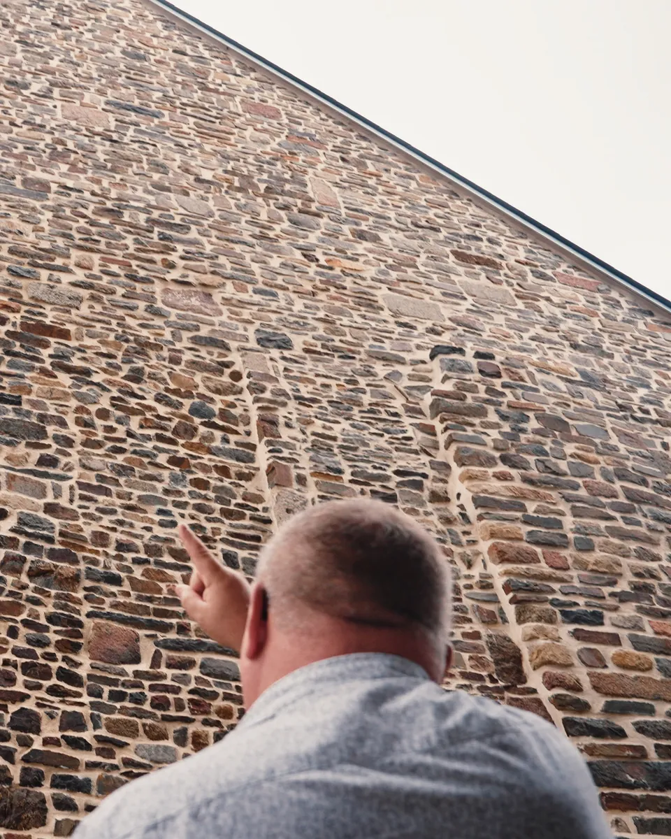 Un homme, vu de dos, montre du doigt le haut d’un grand pignon en moellons rejointoyés.