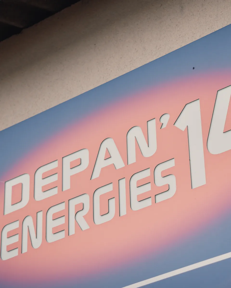 L’enseigne Depan’energies 14, vue de biais : lettres blanches découpées sur un panneau bleu au centre rose, fixé au mur crépi d’un bâtiment.