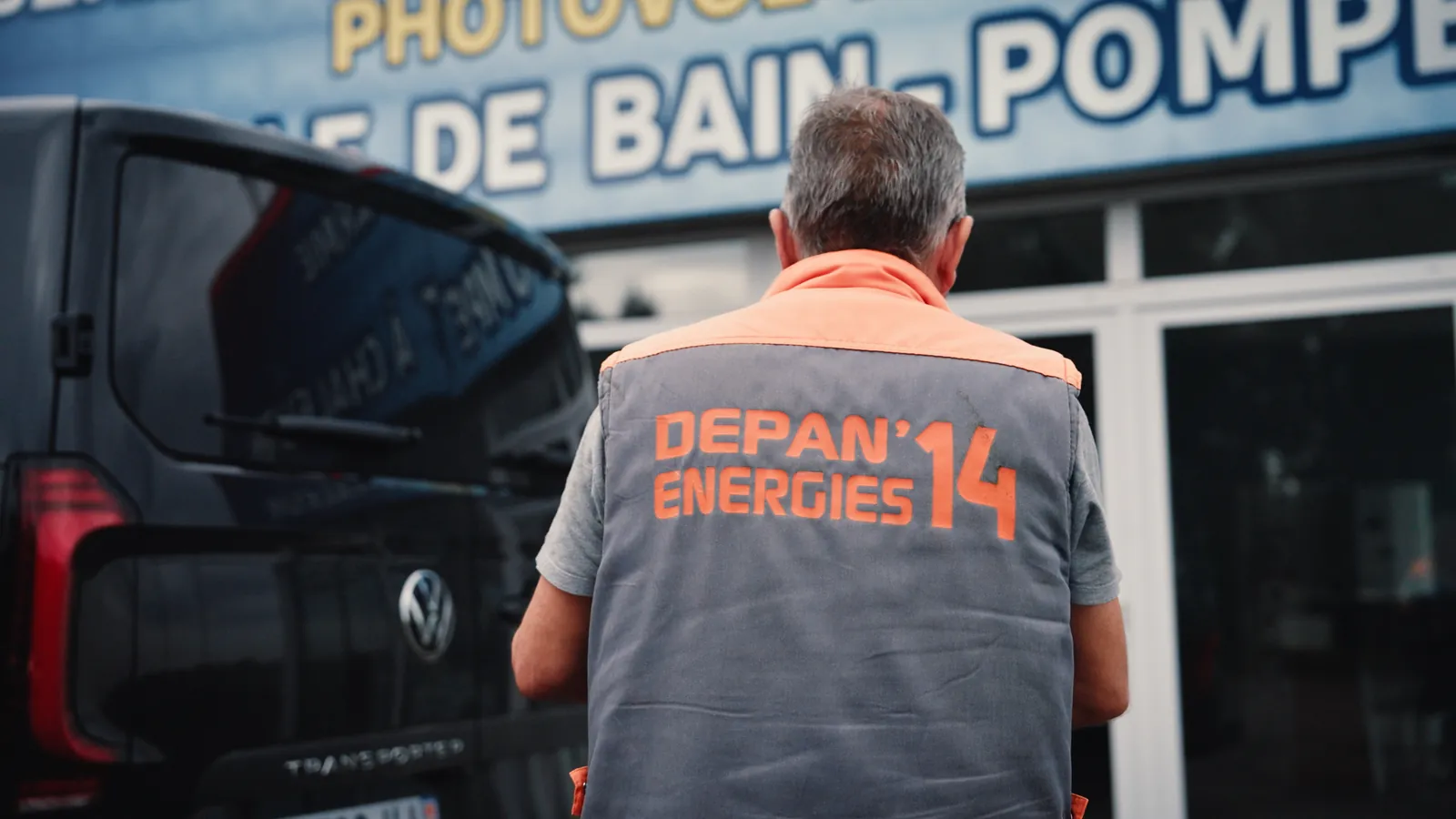 Un technicien aux cheveux gris, vu de dos, en gilet gris et orange floqué Depan’energies 14, devant un utilitaire noir et une grande enseigne bleue.