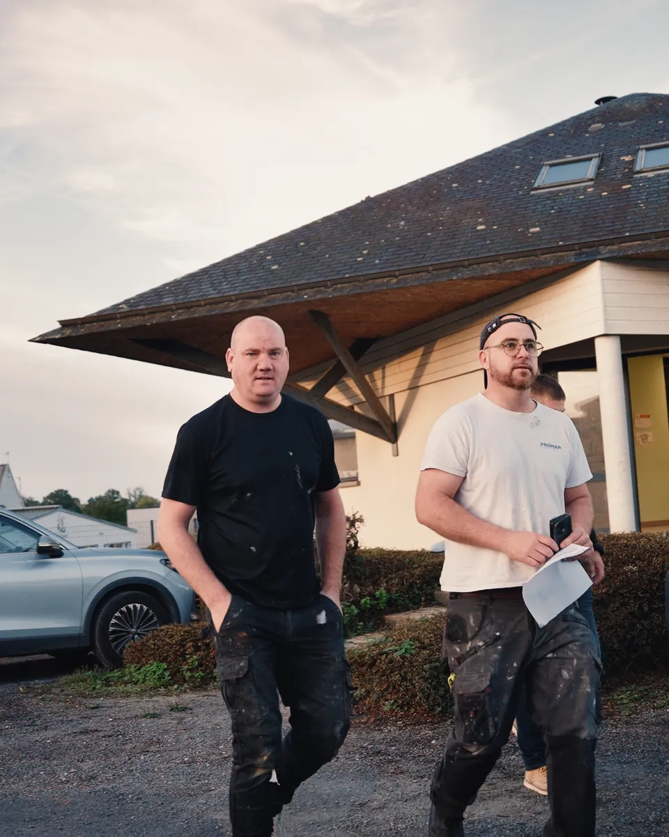 Deux techniciens de Deperiers marchent vers l’objectif devant un bâtiment à toit d’ardoise : l’un en tee-shirt noir, l’autre en tee-shirt blanc, des feuilles à la main.