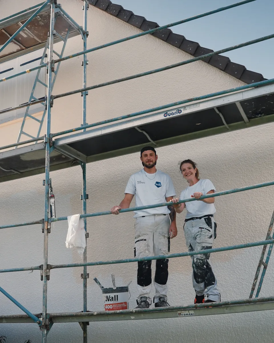Deux peintres en tenue blanche, un homme et une femme, posent debout sur l’échafaudage d’un pignon, les bras sur le garde-corps.