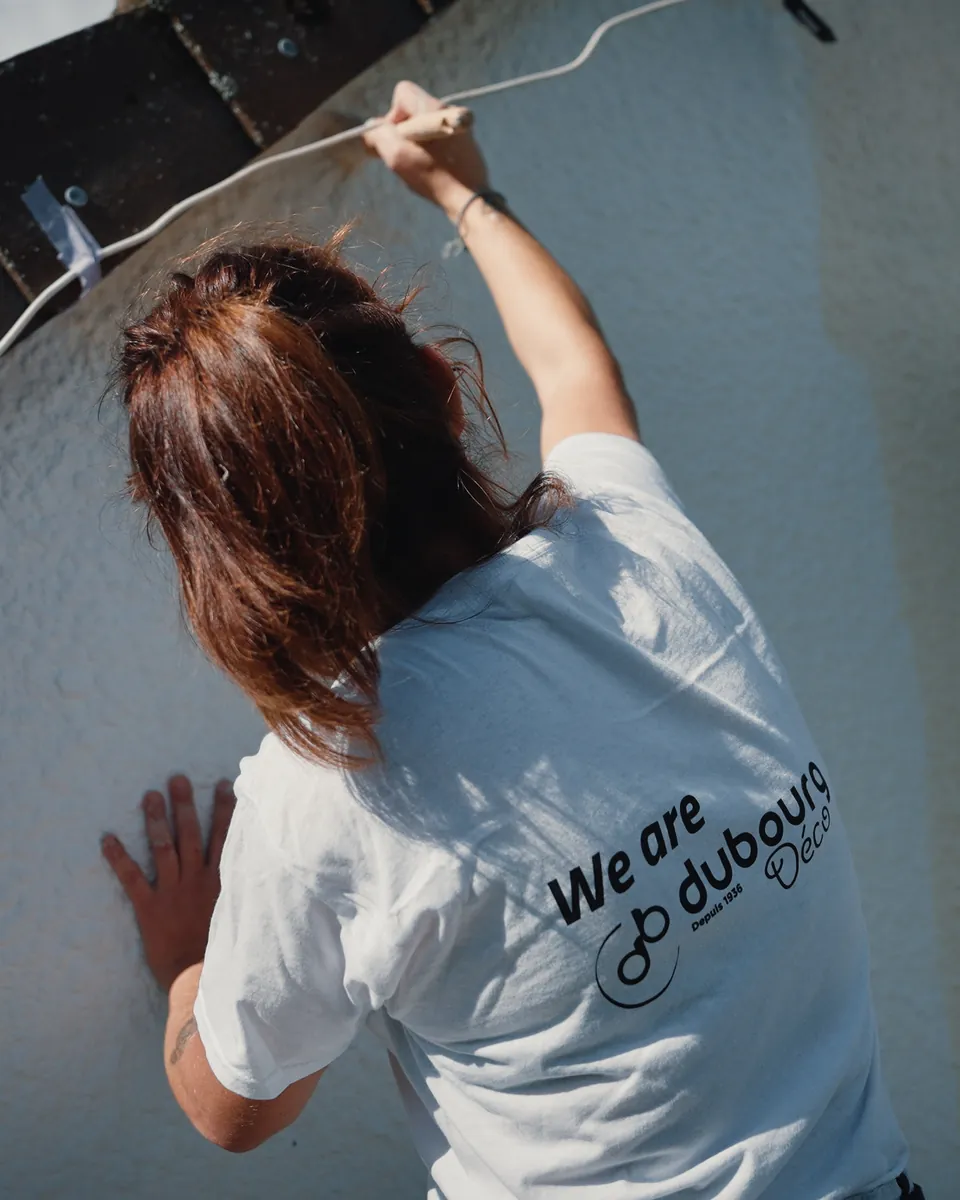 Une peintre, de dos, en tee-shirt Dubourg Déco, reprend la façade au pinceau sous la rive du toit, à côté d’un montant d’échafaudage et d’un volet.