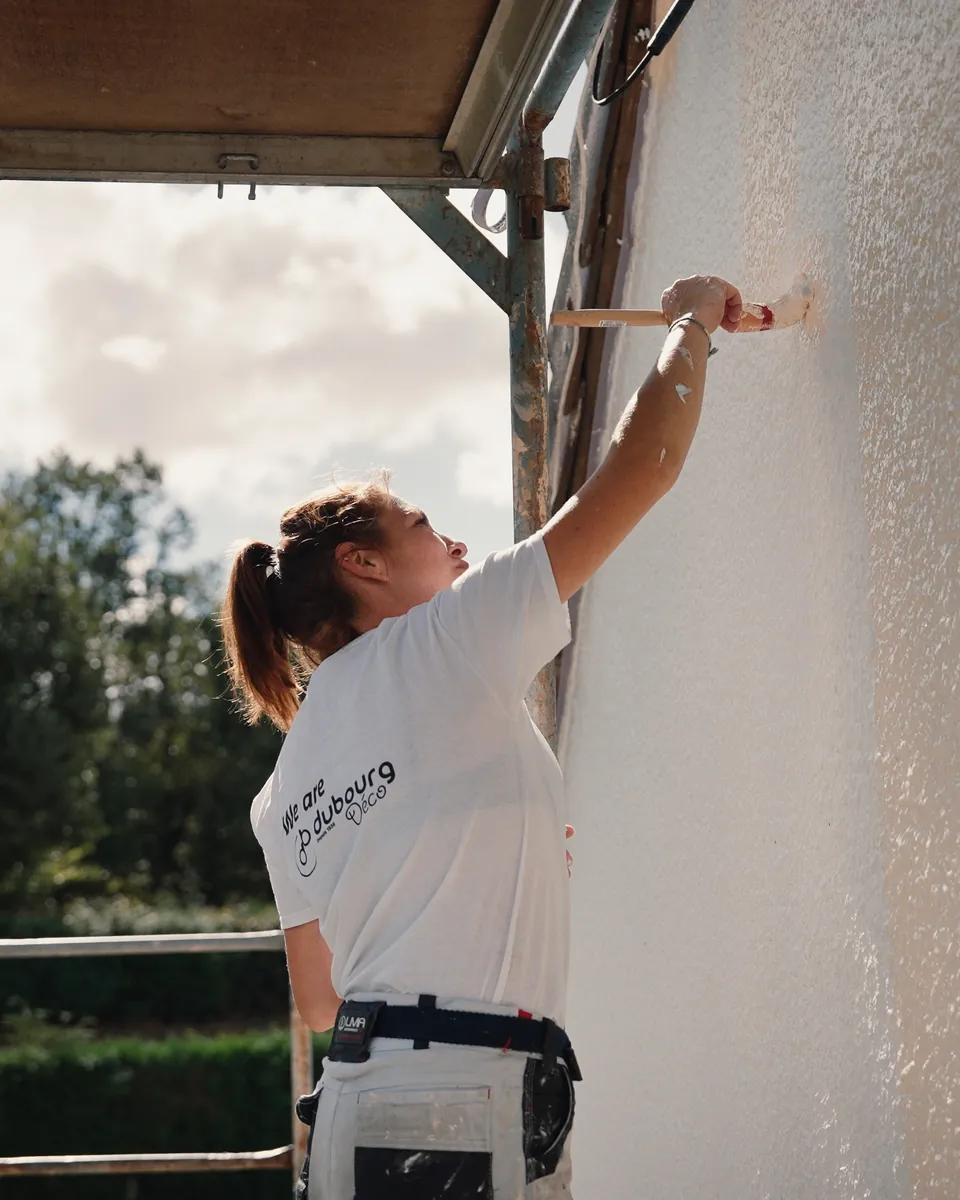 Une peintre en tee-shirt Dubourg Déco, bras levé, passe un pinceau sur un crépi blanc depuis l’échafaudage.