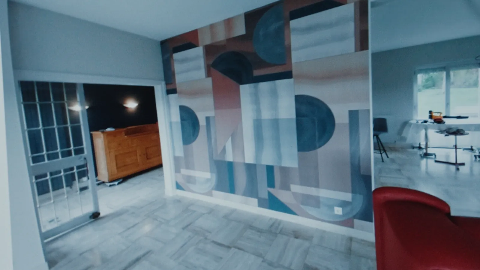 Photographie d’un intérieur affichée sur un écran : un grand panneau mural à motifs géométriques bleus, roses et rouille, une porte vitrée à petits carreaux, un buffet en bois et un sol clair.