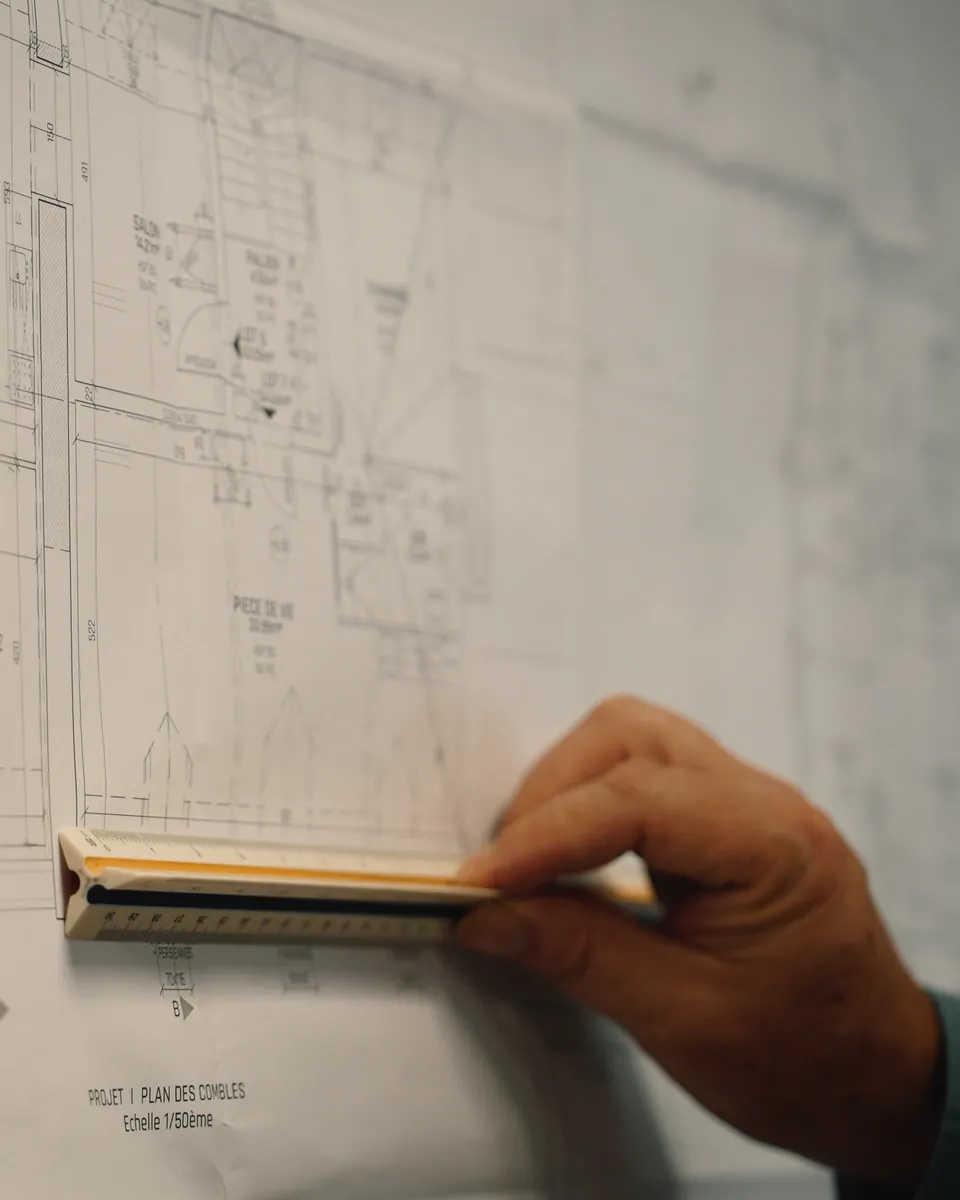 Une main pose une règle graduée sur un plan à l’échelle 1/50e intitulé « Plan des combles », où l’on lit salon, palier et pièce de vie.