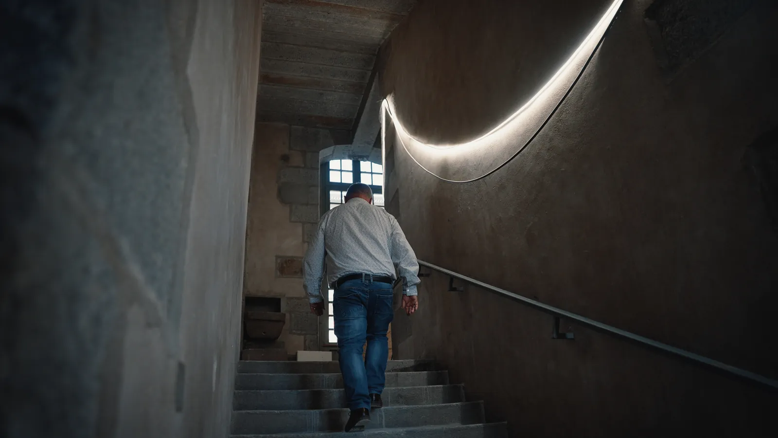 Cage d’escalier rénovée, paroi courbe éclairée par un ruban de LED devant un mur de pierre.