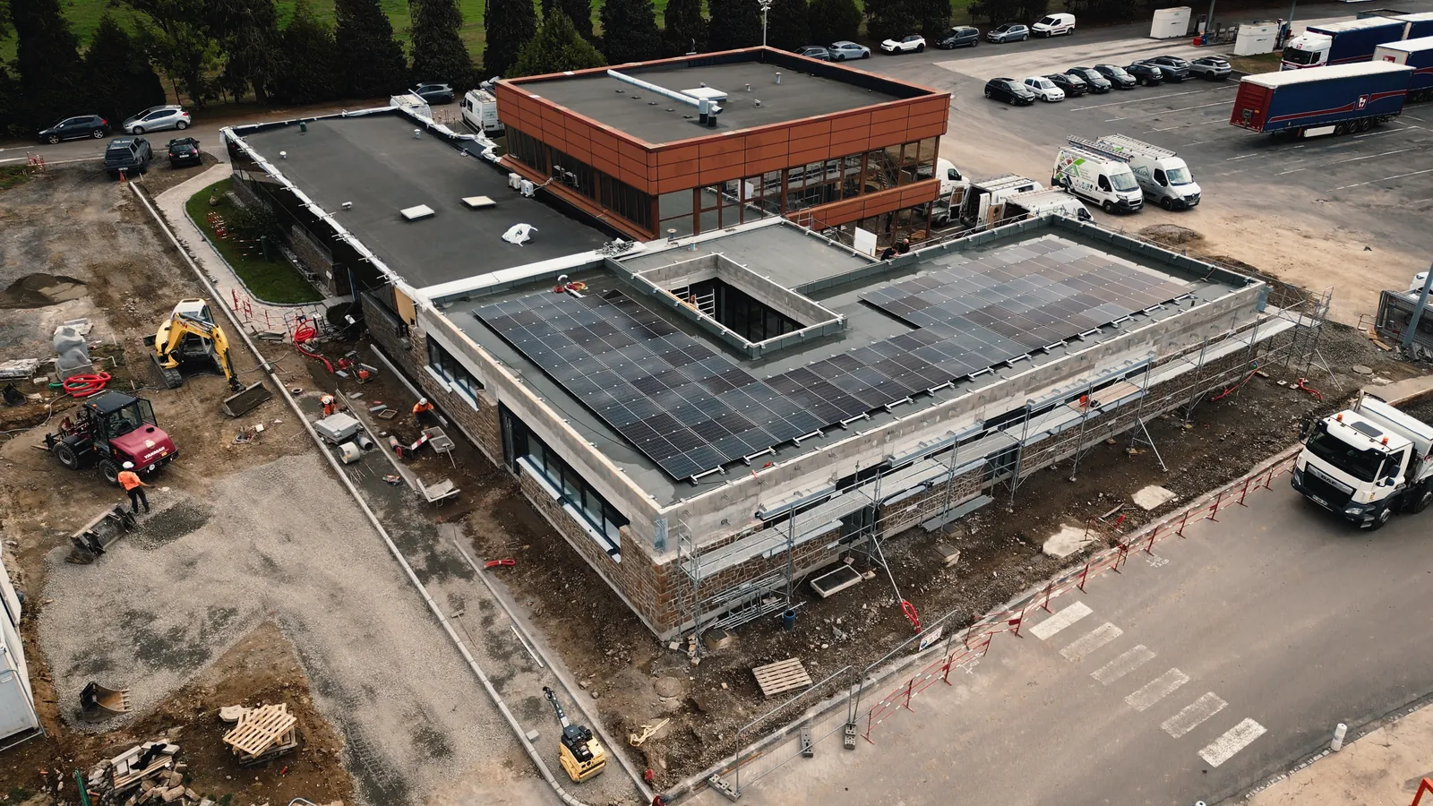 Vue du ciel d’un chantier de gros œuvre : bâtiment aux murs de pierre et toiture en panneaux solaires, engins de terrassement autour.