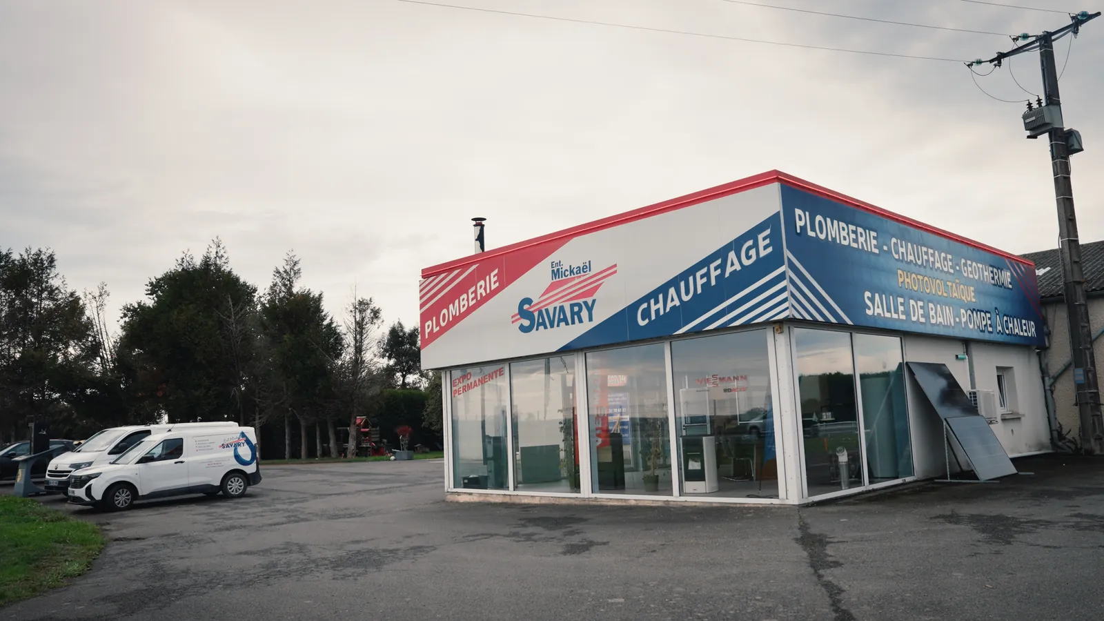 Le showroom de SAVARY : un bâtiment d’angle à façade vitrée, sous une enseigne qui annonce plomberie, chauffage, géothermie, photovoltaïque, salle de bain et pompe à chaleur.