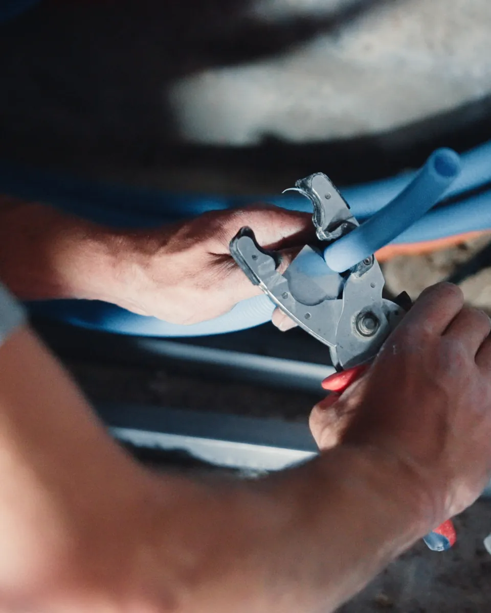 Gros plan de deux mains qui coupent un tube de plomberie bleu avec un outil de coupe, près d’un collecteur.